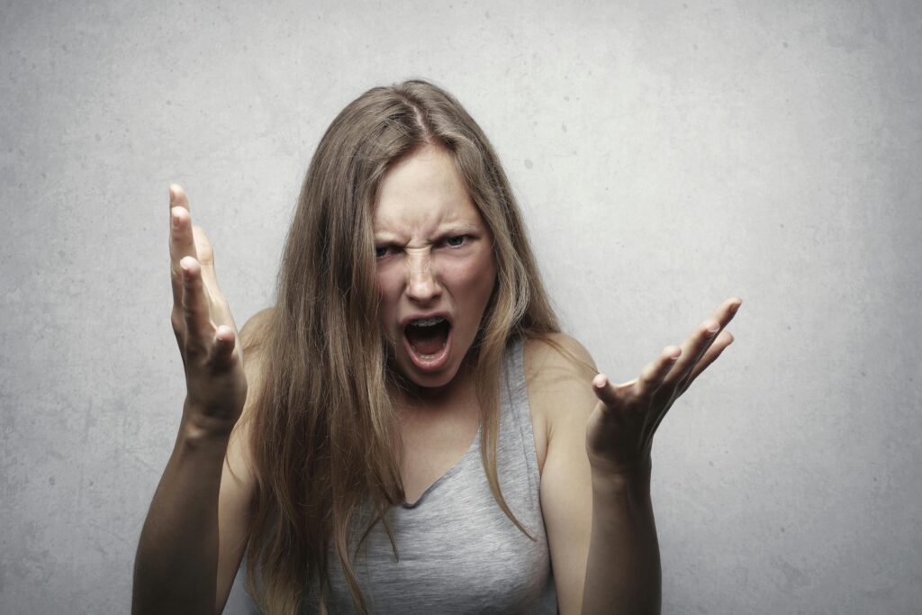 A young woman showing intense anger and frustration, yelling with hands raised against a gray background.